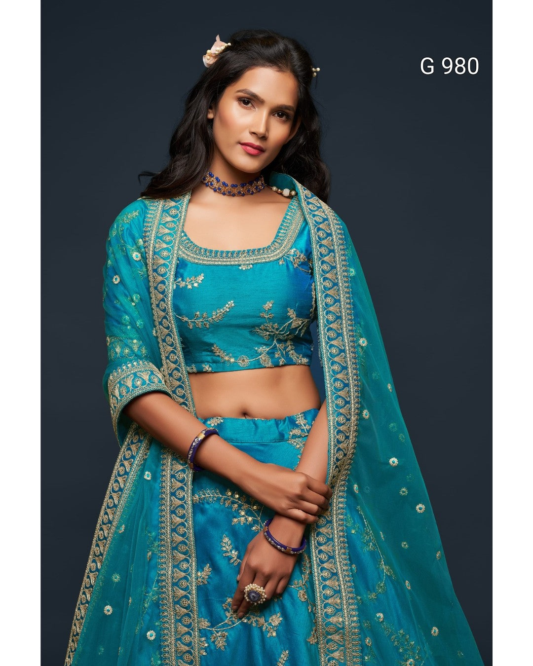 A woman modeling a teal blue traditional Indian outfit with gold embroidery, featuring a crop top, flared skirt, and matching dupatta, set against a dark gray studio background.