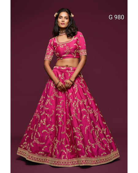 A woman modeling a bright pink traditional Indian lehenga choli set with gold floral embroidery, set against a solid dark maroon background. The text 'G 980' is visible in the top right corner.