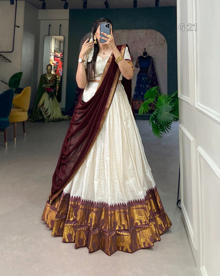 Person wearing a white saree with maroon and gold border in a room.