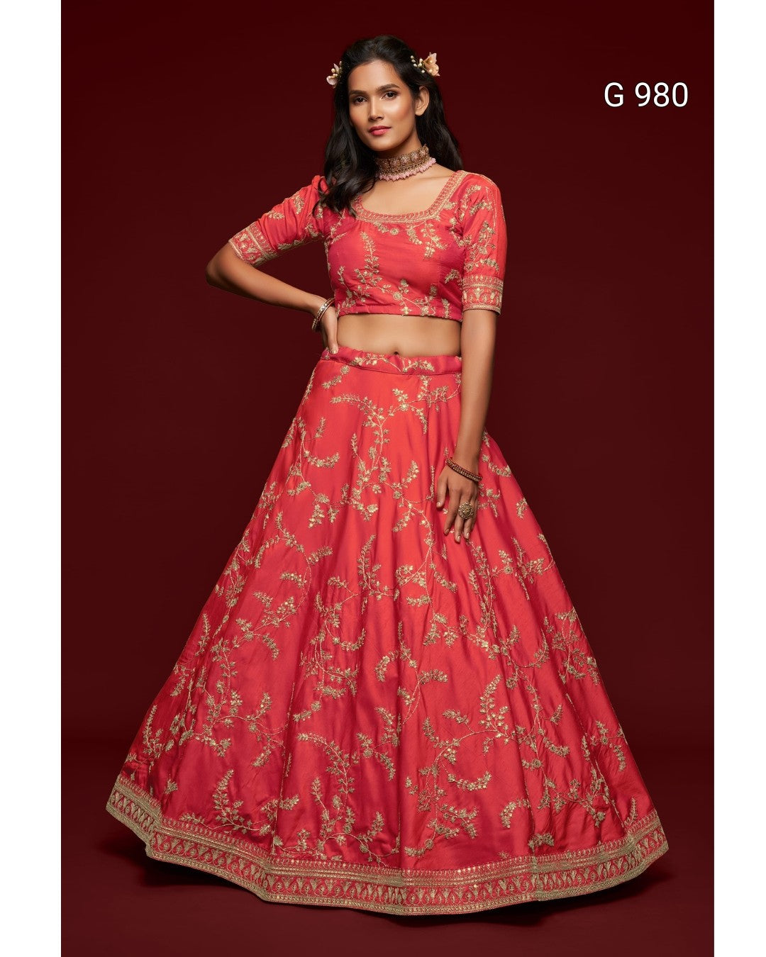 A woman models a traditional Indian lehenga choli set in coral pink with gold floral embroidery, set against a solid dark red background. The text 'G 980' is visible in the top right corner.