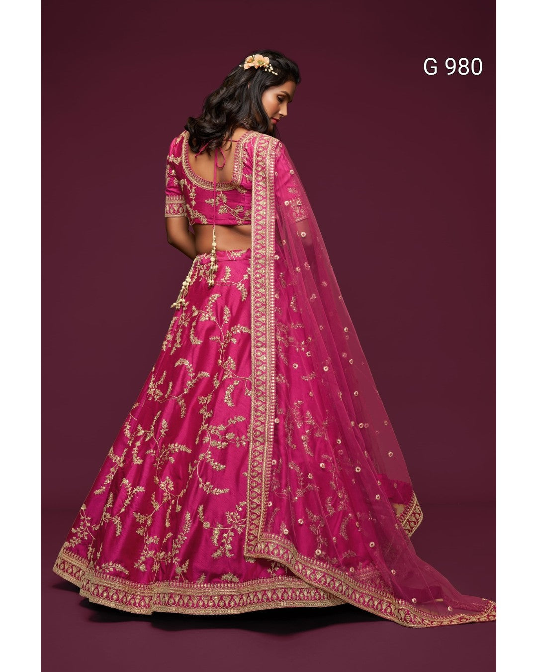 A woman wearing a bright pink embroidered Indian lehenga choli suit with a matching dupatta, shown from the back against a dark maroon studio background. The outfit has intricate gold floral embroidery and a decorative border.
