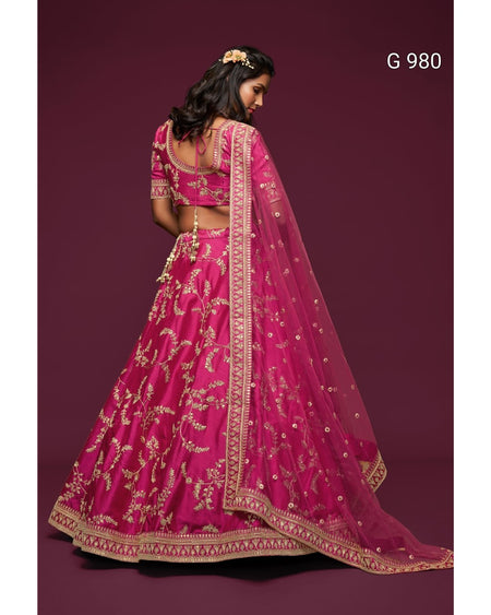 A woman wearing a bright pink embroidered Indian lehenga choli suit with a matching dupatta, shown from the back against a dark maroon studio background. The outfit has intricate gold floral embroidery and a decorative border.