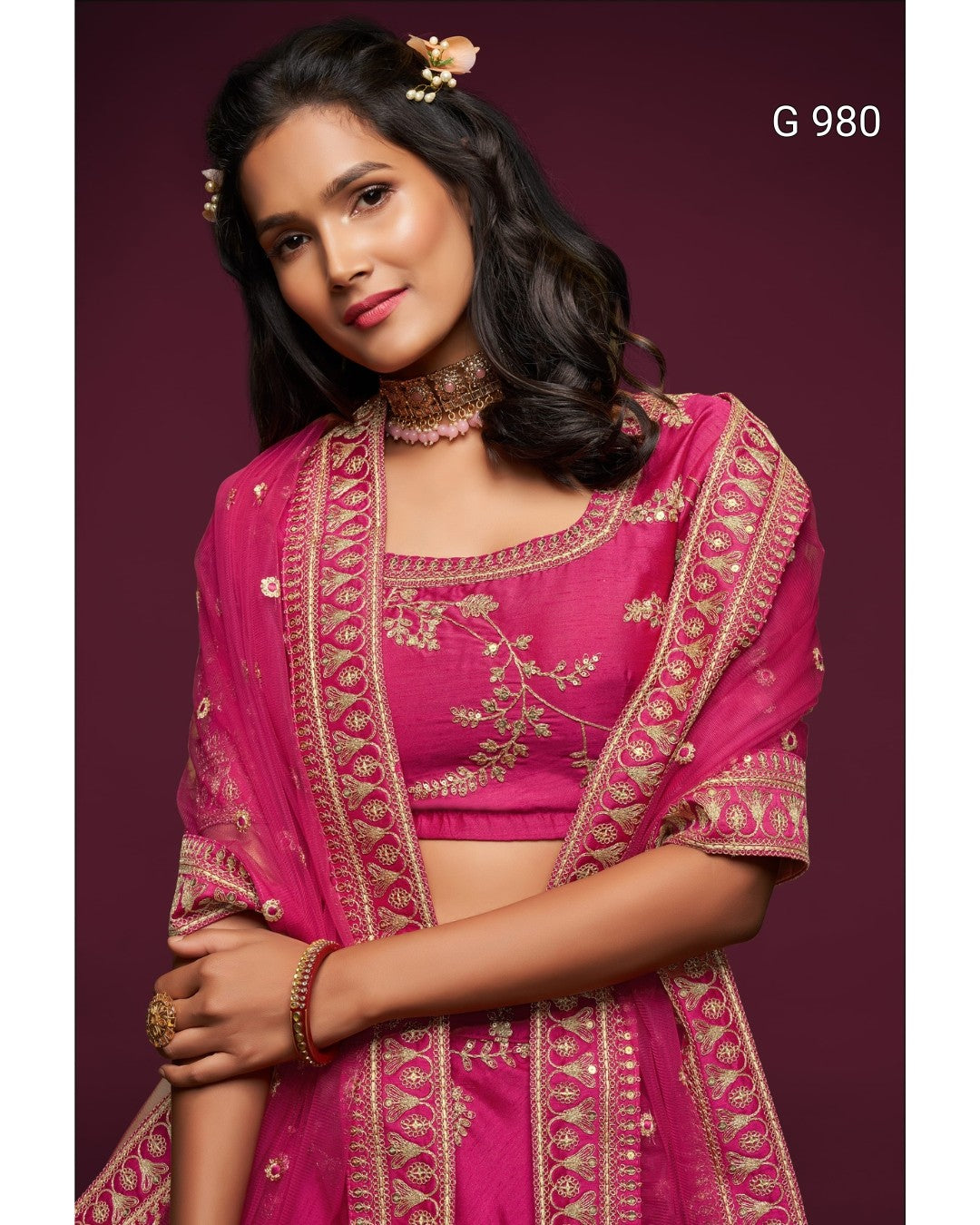 A woman modeling a bright pink traditional Indian outfit with gold embroidery and a matching sheer dupatta, posed against a dark burgundy studio background. The text 'G 980' appears in the top right corner.
