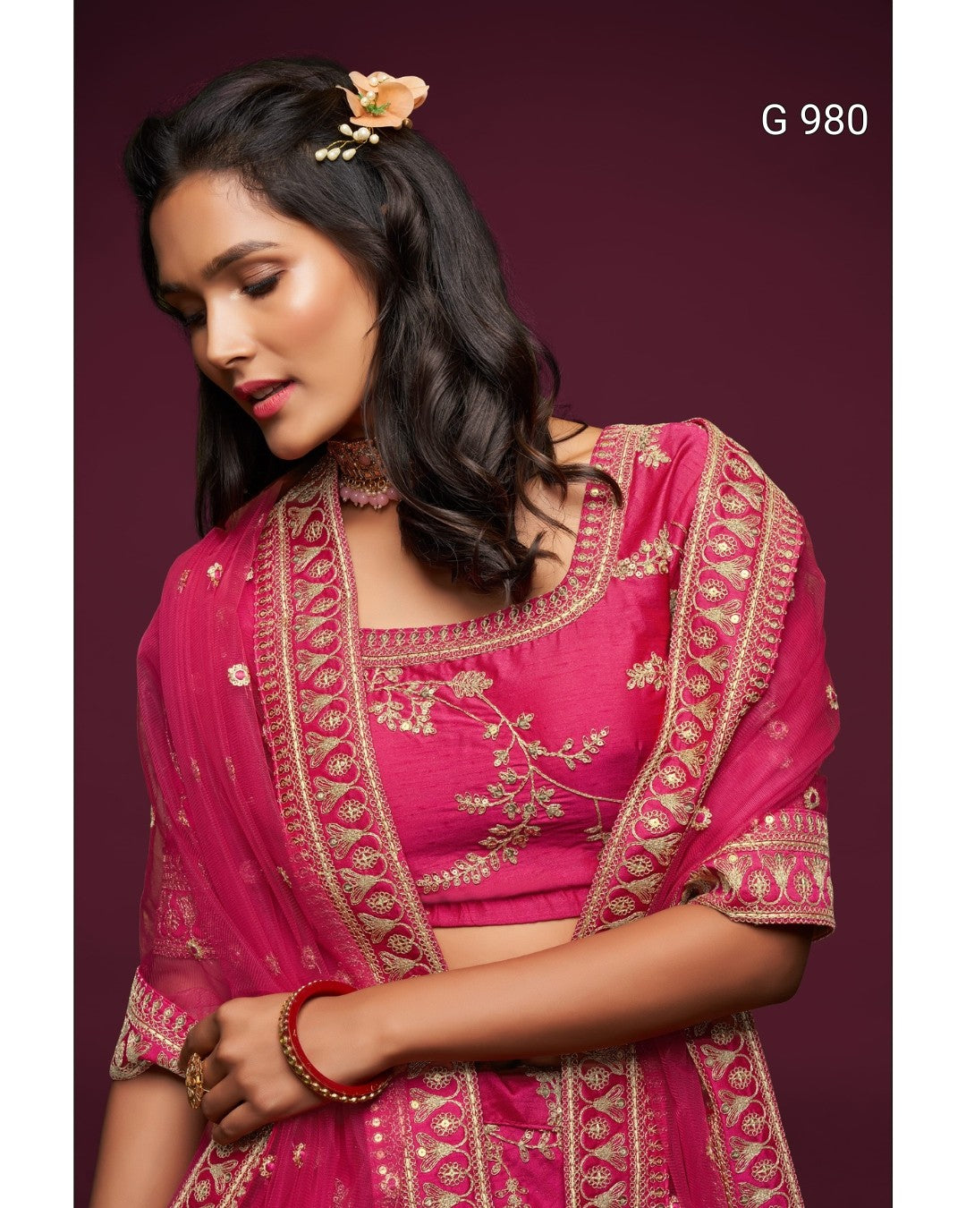 A woman wearing a bright pink traditional outfit with ornate gold embroidery and a matching sheer dupatta, posed against a dark burgundy background. The text 'G 980' appears in the top right corner.