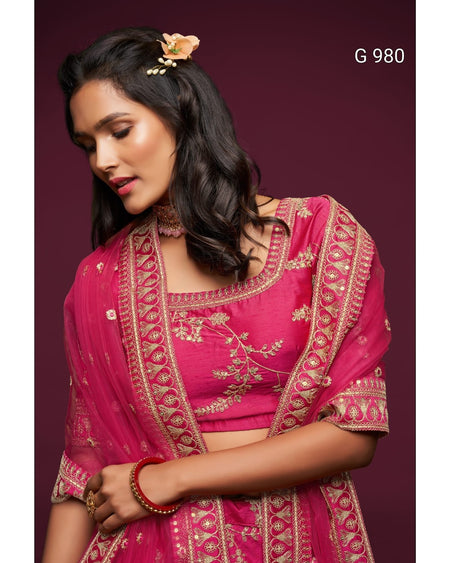 A woman wearing a bright pink traditional outfit with ornate gold embroidery and a matching sheer dupatta, posed against a dark burgundy background. The text 'G 980' appears in the top right corner.