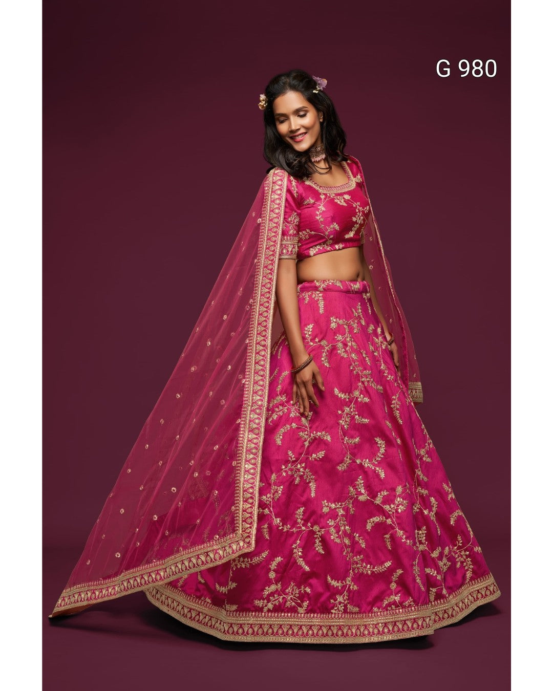 A woman modeling a bright pink lehenga choli set with gold floral embroidery and a matching sheer dupatta, posed against a solid dark maroon background. The text 'G 980' is visible in the top right corner.