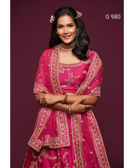 A woman modeling a bright pink traditional Indian outfit with gold embroidery, featuring a flared skirt, embroidered blouse, and matching dupatta, set against a solid dark maroon background.