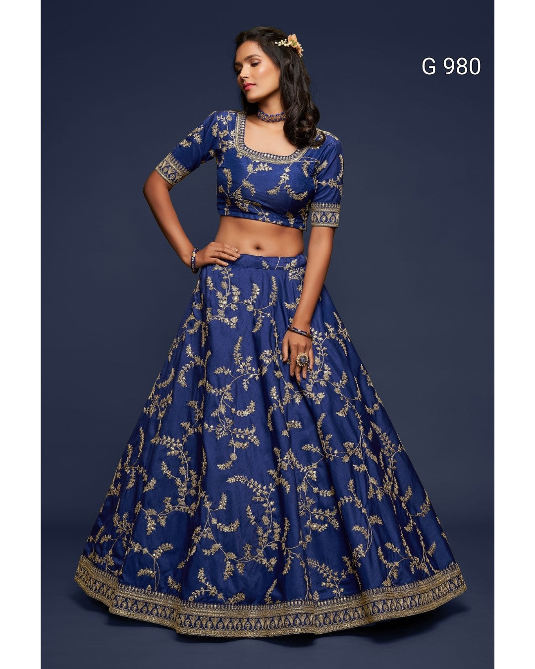 A woman modeling a royal blue lehenga choli set with intricate gold floral embroidery, set against a dark blue studio background. The text 'G 980' is visible in the top right corner.