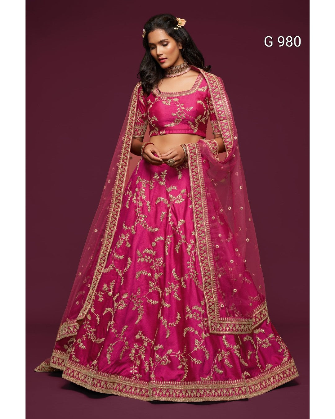 A woman modeling a bright pink traditional Indian lehenga choli set with gold floral embroidery and a matching sheer dupatta, set against a solid dark maroon background. The text 'G 980' is visible in the top right corner.