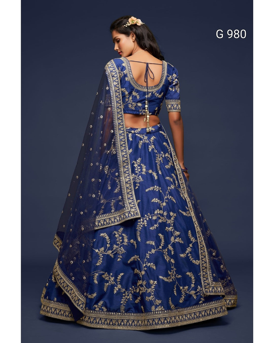A woman modeling a royal blue lehenga choli with intricate gold embroidery, shown from the back against a dark blue studio background. The text 'G 980' is visible in the top right corner.