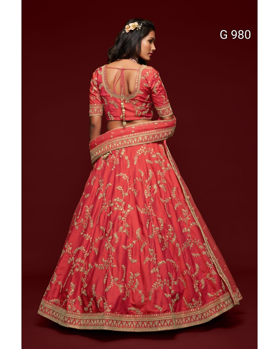 A woman modeling a red and gold embroidered Indian lehenga choli suit from the back, set against a solid dark red background. The text 'G 980' is visible in the top right corner.