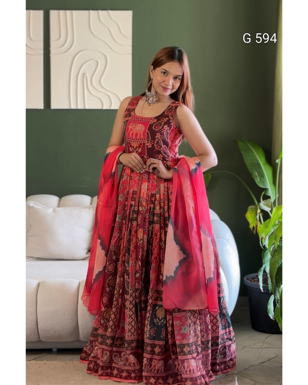 A woman models a traditional Indian ethnic outfit consisting of a red and black patterned lehenga choli with a matching sheer dupatta, set against a dark green wall and a white sofa.