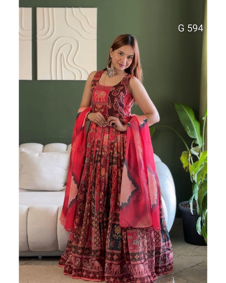A woman models a traditional Indian ethnic outfit consisting of a red and black patterned lehenga choli with a matching sheer dupatta, set against a dark green wall and a white sofa.