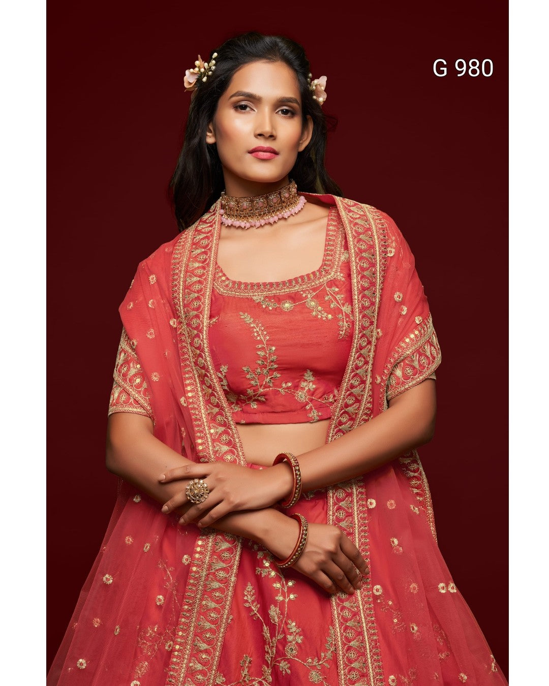 A woman modeling a coral pink embroidered traditional Indian outfit with a matching dupatta, set against a solid dark red background. The outfit features intricate gold embroidery and has the text 'G 980' in the top right corner.