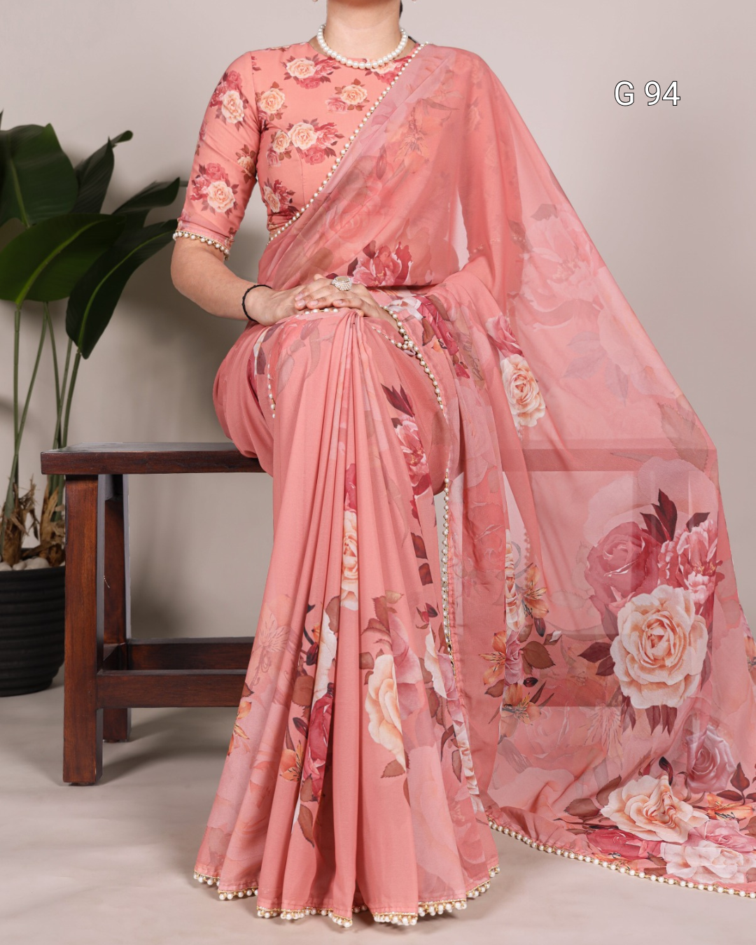 Woman wearing a pink floral saree with a matching blouse, sitting on a wooden stool.