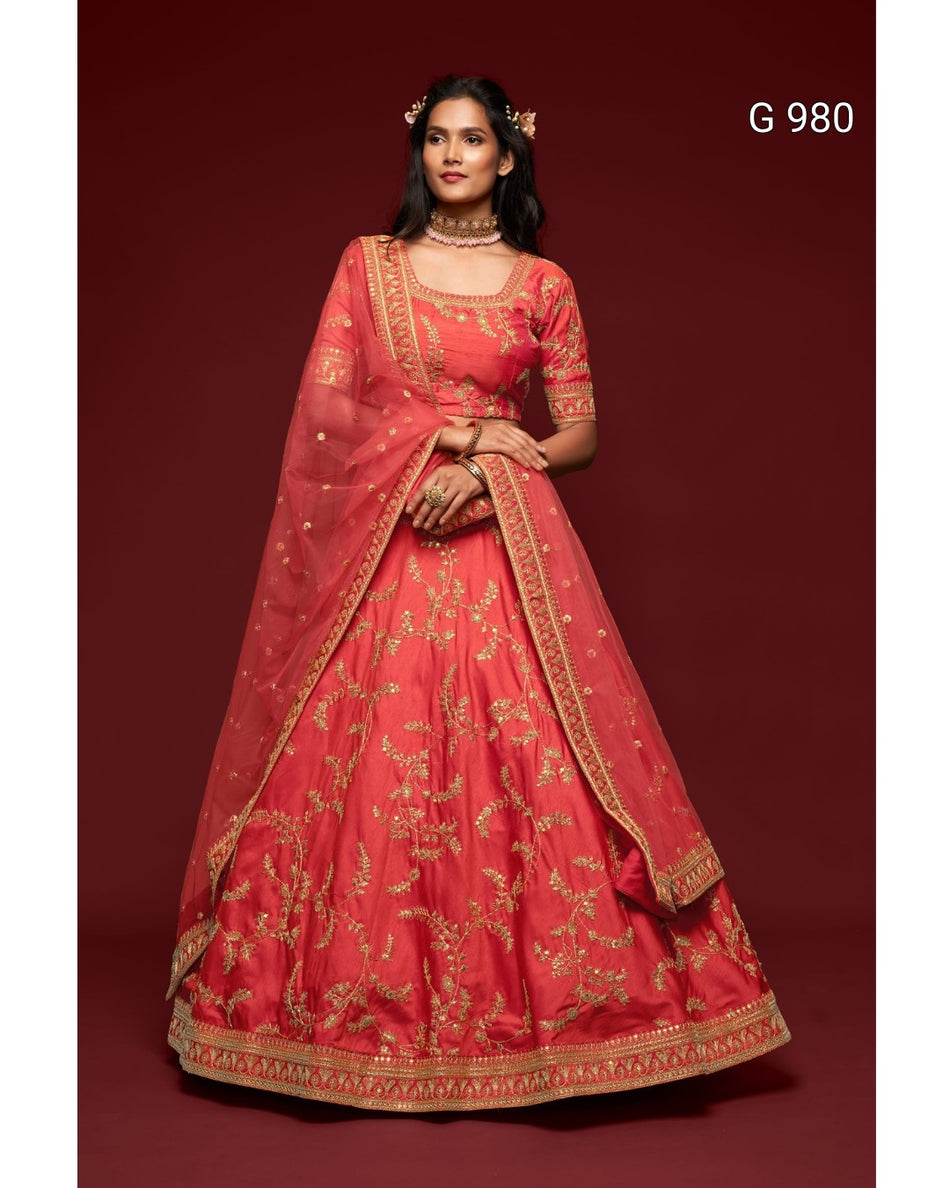 A woman modeling a traditional red lehenga choli set with intricate gold embroidery, set against a solid dark red background. The set includes a red flared skirt (lehenga), a matching embroidered blouse (choli), and a sheer red dupatta with gold borders.
