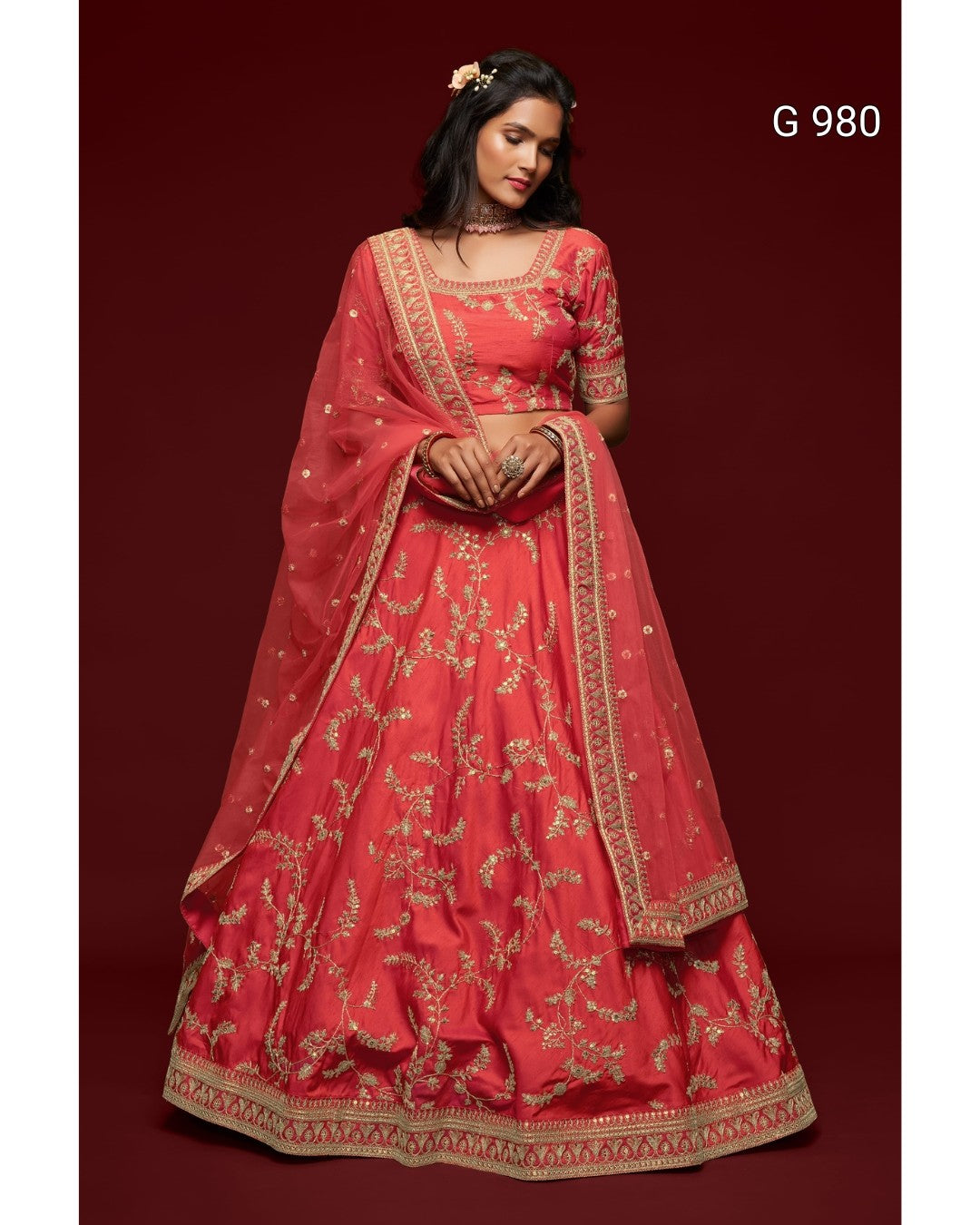 A woman modeling a traditional red lehenga choli set with gold embroidery, featuring a red skirt, matching blouse, and dupatta, set against a dark red background with the text 'G 980' in the top right.