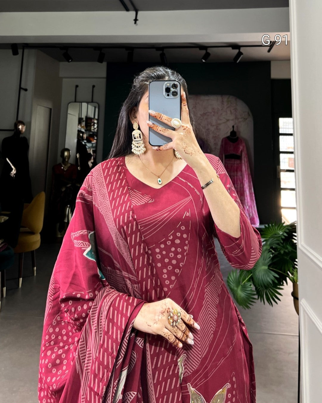 Woman taking a mirror selfie wearing a red patterned dress in a room with plants and furniture.