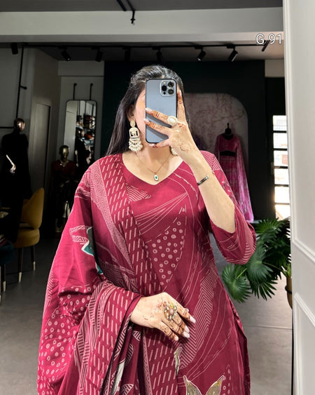 Woman taking a mirror selfie wearing a red patterned dress in a room with plants and furniture.