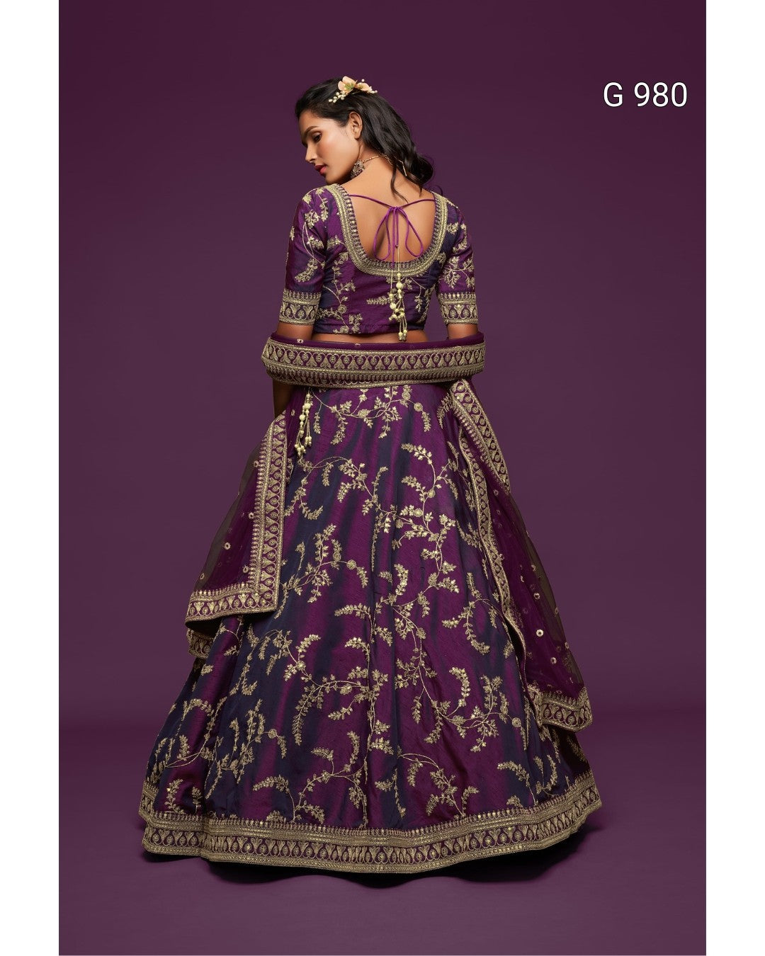 A woman modeling a traditional Indian lehenga choli set from the back, featuring a deep purple fabric with intricate gold floral embroidery and a matching dupatta, set against a solid purple background.