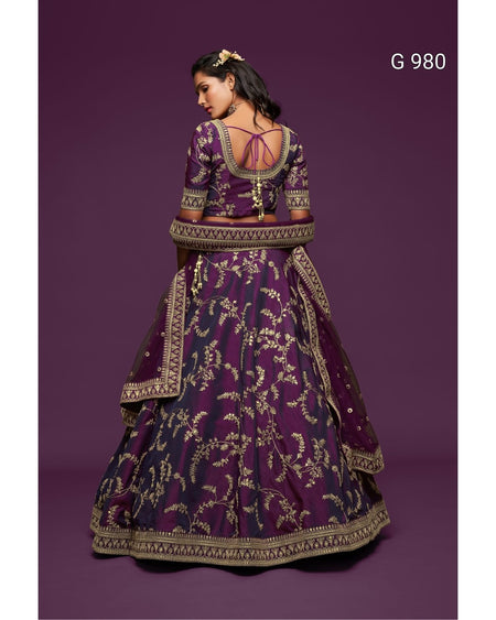A woman modeling a traditional Indian lehenga choli set from the back, featuring a deep purple fabric with intricate gold floral embroidery and a matching dupatta, set against a solid purple background.