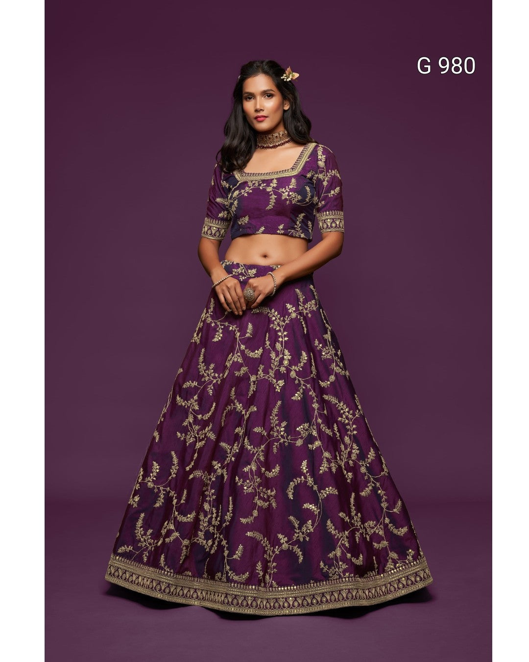 A woman modeling a deep purple lehenga choli set with intricate gold embroidery, set against a solid dark purple studio background. The text 'G 980' is visible in the top right corner.