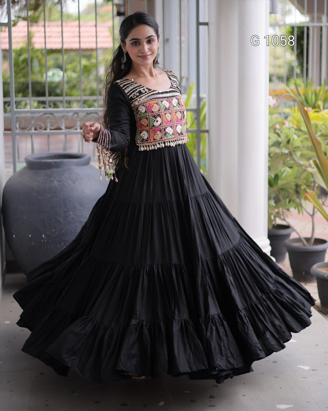 A woman wearing a black tiered Anarkali gown with a heavily embroidered multicolor bodice, standing in a courtyard setting.