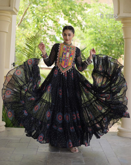 A woman wearing a black traditional long dress with colorful embroidered neckline and circular patterned details, holding out her voluminous skirt in a courtyard setting.