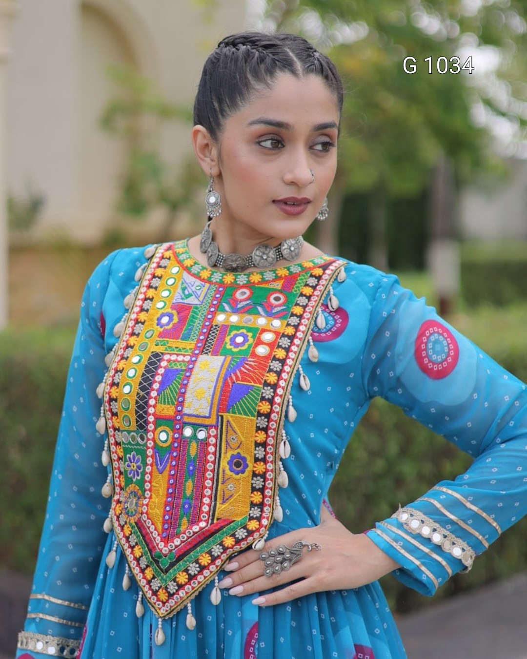 A woman wearing a bright blue traditional embroidered outfit with a heavily decorated multicolored embroidered chest panel, silver jewelry, and a blurred outdoor background.