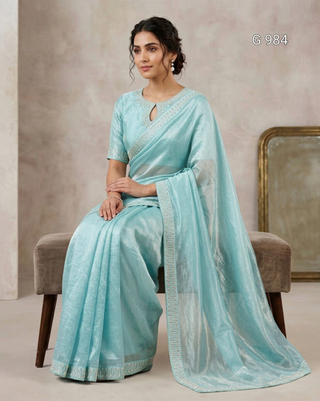 A woman seated on a upholstered bench wearing a light aqua blue saree with a matching sheer drape and embroidered border. The setting is indoors against a textured neutral wall, with a mirror partially visible behind her.