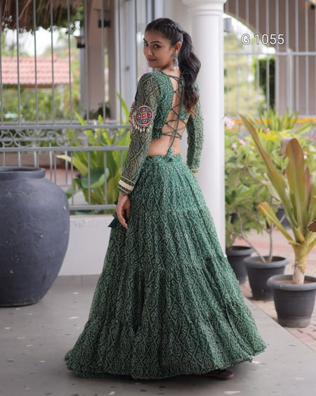 A woman wearing a green embroidered traditional outfit with a long lace skirt and matching blouse, standing on a porch with potted plants in the background.