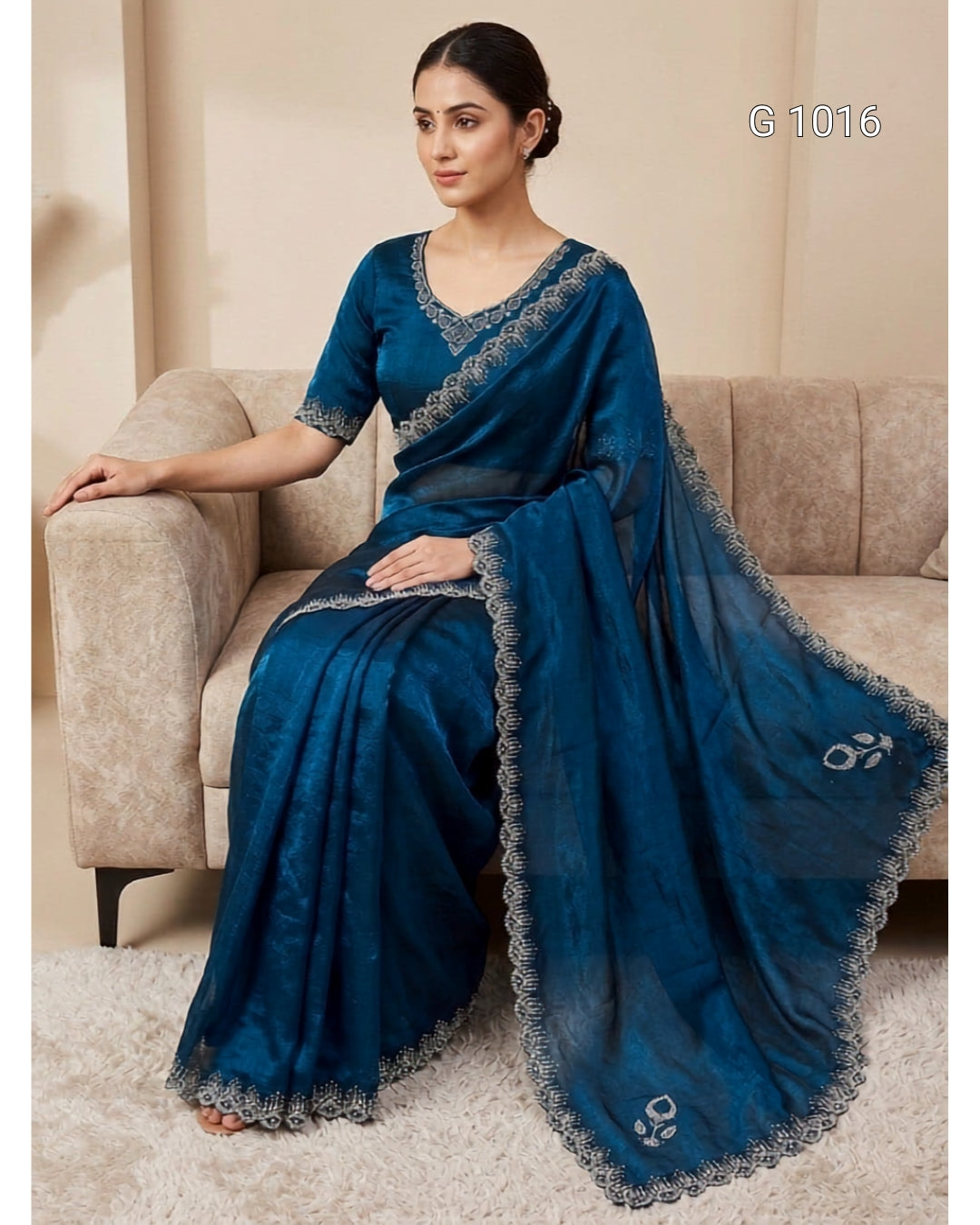 A woman seated on a beige sofa wearing a dark teal blue saree with silver embroidered borders and a matching blouse. The image includes the text 'G 1016' in the top right corner.