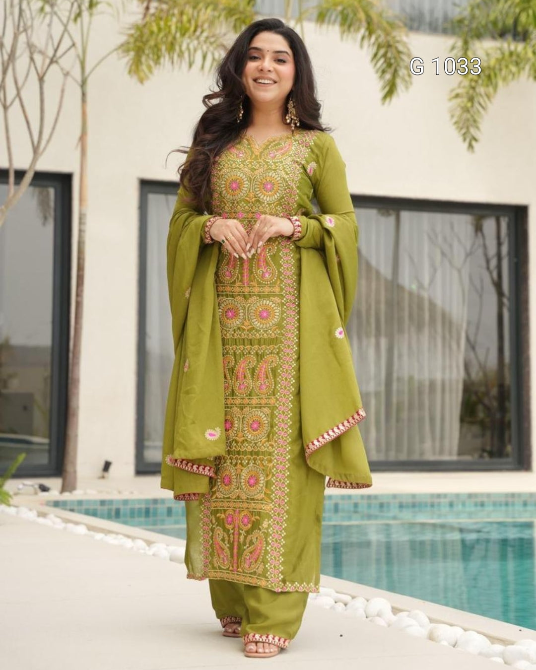 A woman models a traditional olive green salwar kameez suit with intricate gold and pink embroidery, standing on a patio next to a swimming pool.