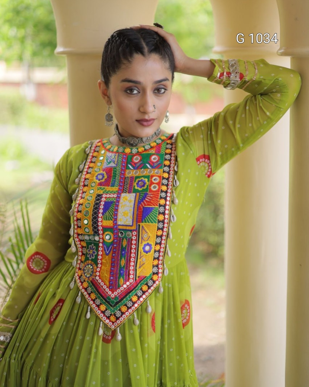 A woman wearing a bright lime green traditional embroidered dress with a heavily decorated multicolor embroidered chest panel, standing in a covered outdoor walkway with cream pillars.