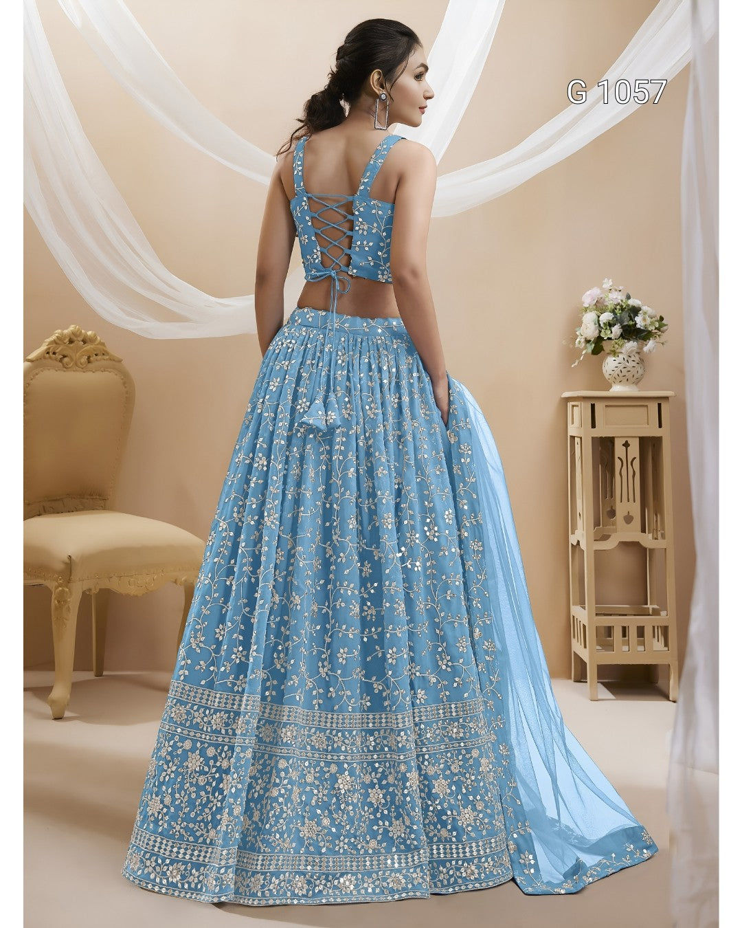 A woman modeling a light blue embroidered lehenga choli set from the back, featuring intricate silver embroidery, a cutout back, and a matching dupatta, set against a warm beige background with draped white fabric.