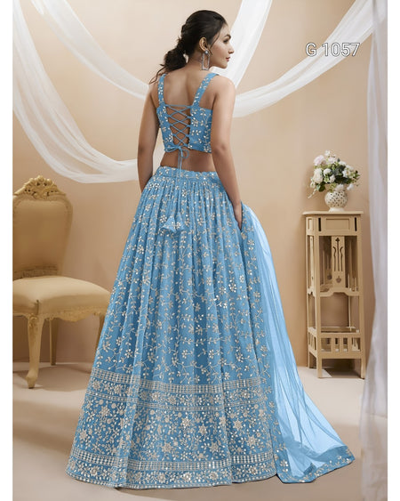 A woman modeling a light blue embroidered lehenga choli set from the back, featuring intricate silver embroidery, a cutout back, and a matching dupatta, set against a warm beige background with draped white fabric.