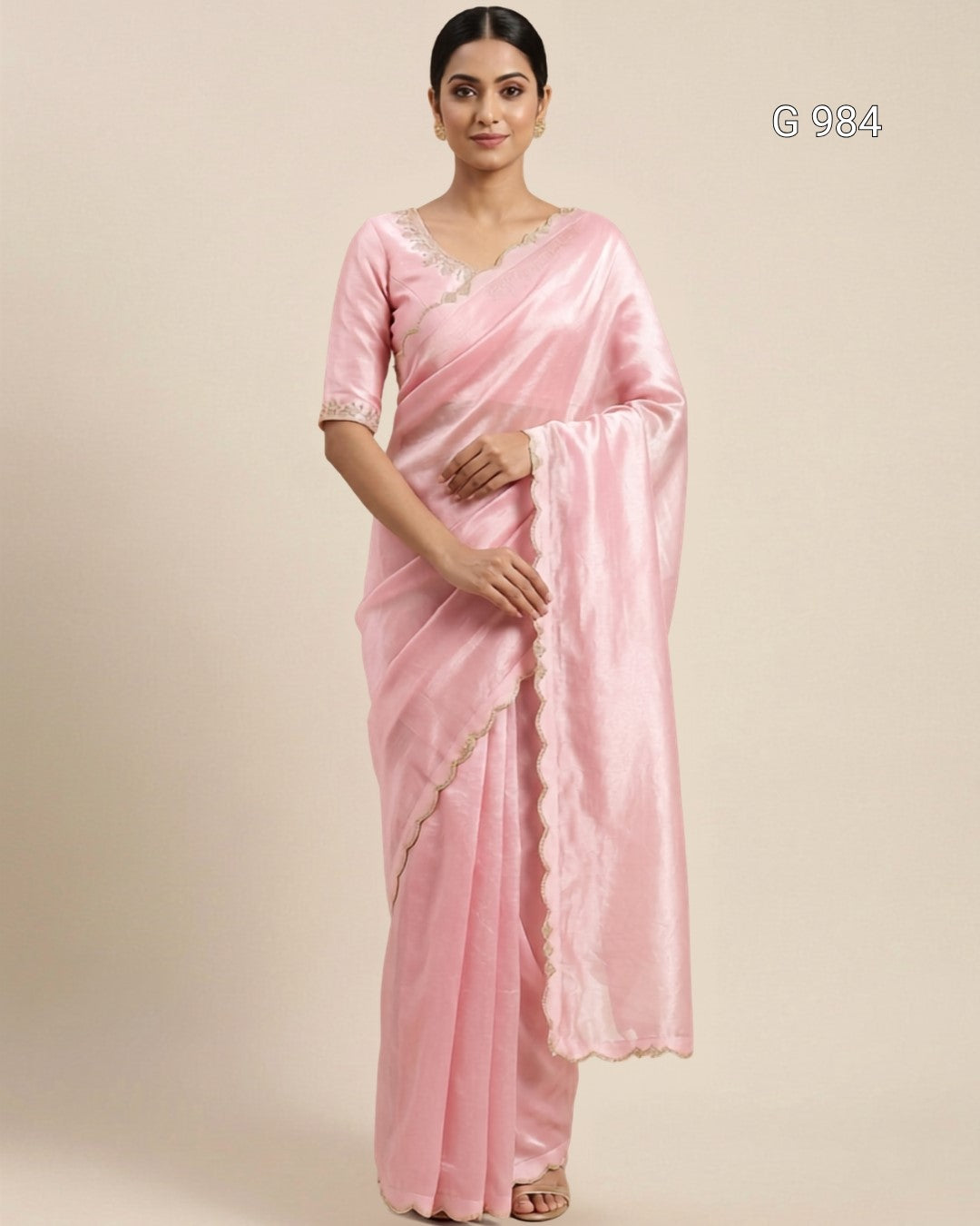 A woman modeling a light pink silk saree with a matching pink blouse, featuring a scalloped gold border and a small embroidered neckline, set against a solid beige background.