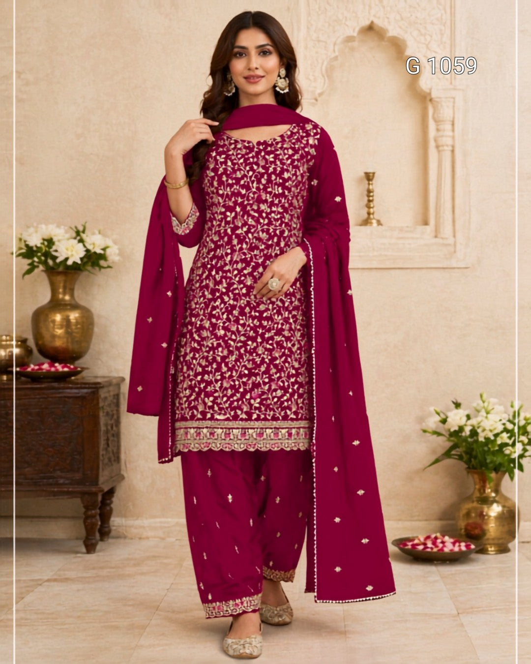 A woman models a deep magenta three-piece ethnic suit with white floral embroidery and gold zari borders, featuring a kurta, pants, and a dupatta, set against a traditional Indian architectural background.