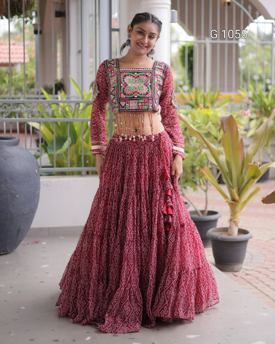 A woman models a traditional Indian lehenga choli outfit in a dusty rose/magenta color with intricate multicolor embroidery on the bodice. The background is a patio with potted plants and columns.