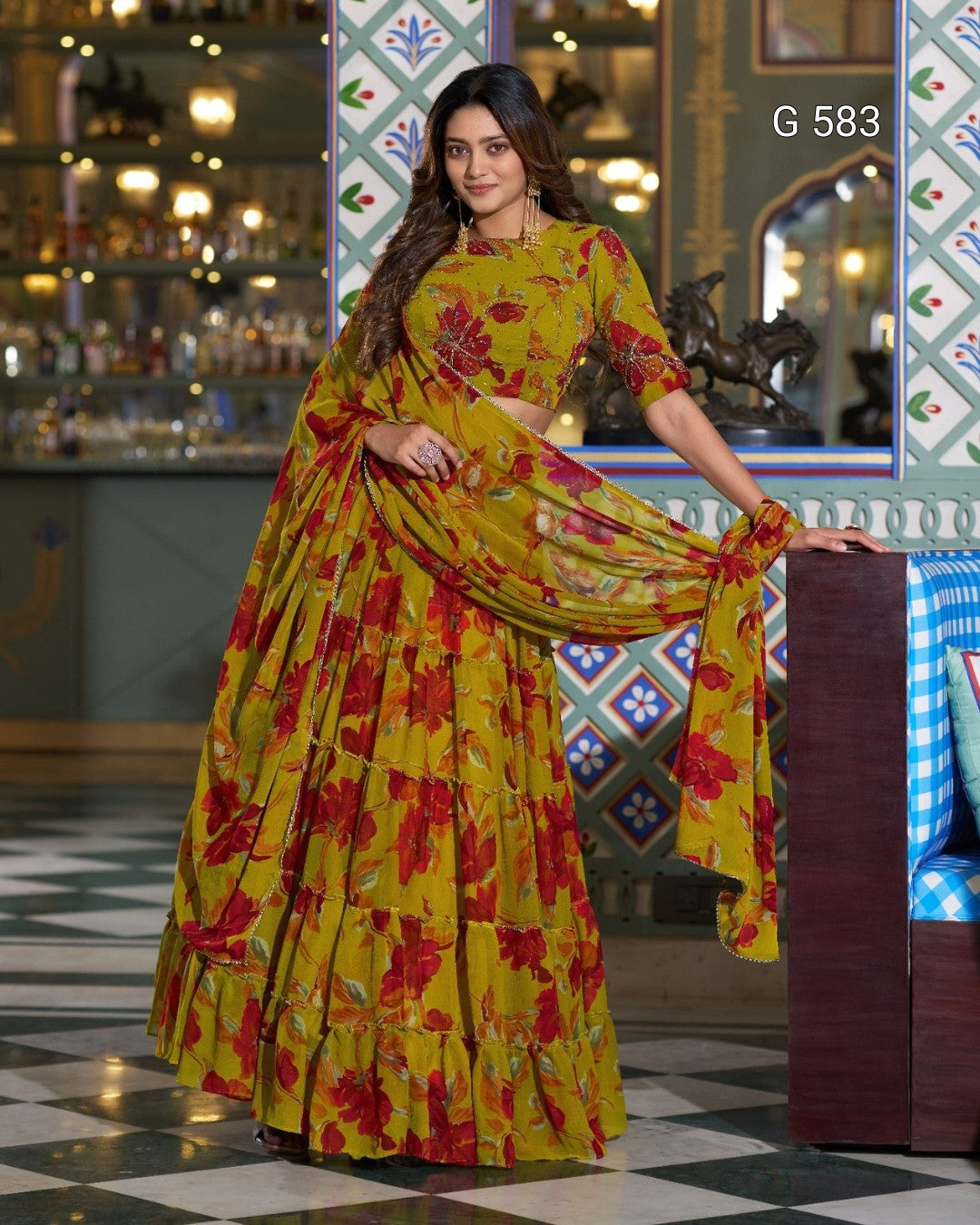 A woman models a yellow and red floral print saree with a matching printed blouse in an indoor setting with patterned walls and a checkered floor.