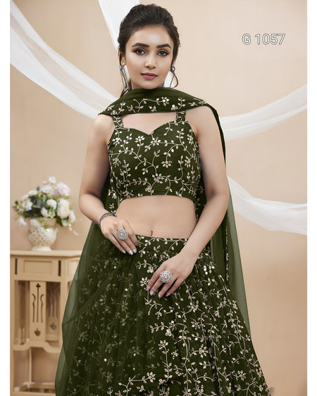 A woman modeling an olive green embroidered lehenga choli set with a matching dupatta, featuring white floral embroidery, set against a beige background with draped white fabric.