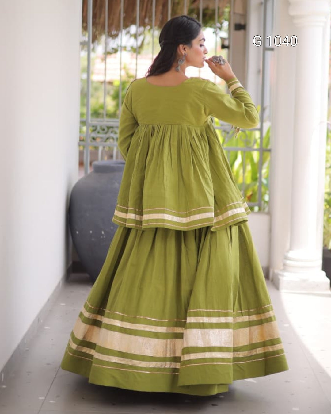 A woman wearing a long-sleeved olive green traditional dress with beige striped borders, shown from the back in an outdoor corridor.