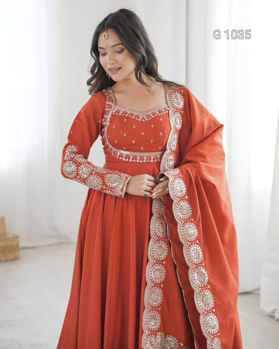 A woman wearing a traditional Indian outfit consisting of a rust-orange Anarkali gown with intricate silver and white embroidery, paired with a matching dupatta draped over her shoulder. The background is a soft, bright white studio setting with sheer curtains.
