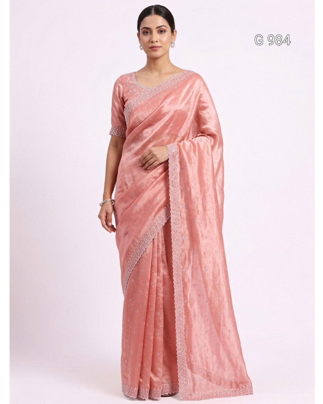 A woman modeling a peach-pink silk saree with a matching short-sleeve blouse. The saree features a subtle sheen and a silver embroidered border, shown against a plain light pink background with the code 'G 904' in the top right corner.