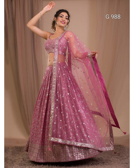 A woman models a pink heavily embellished Indian lehenga choli suit with silver mirror work and a matching sheer dupatta, posed against a beige draped backdrop.