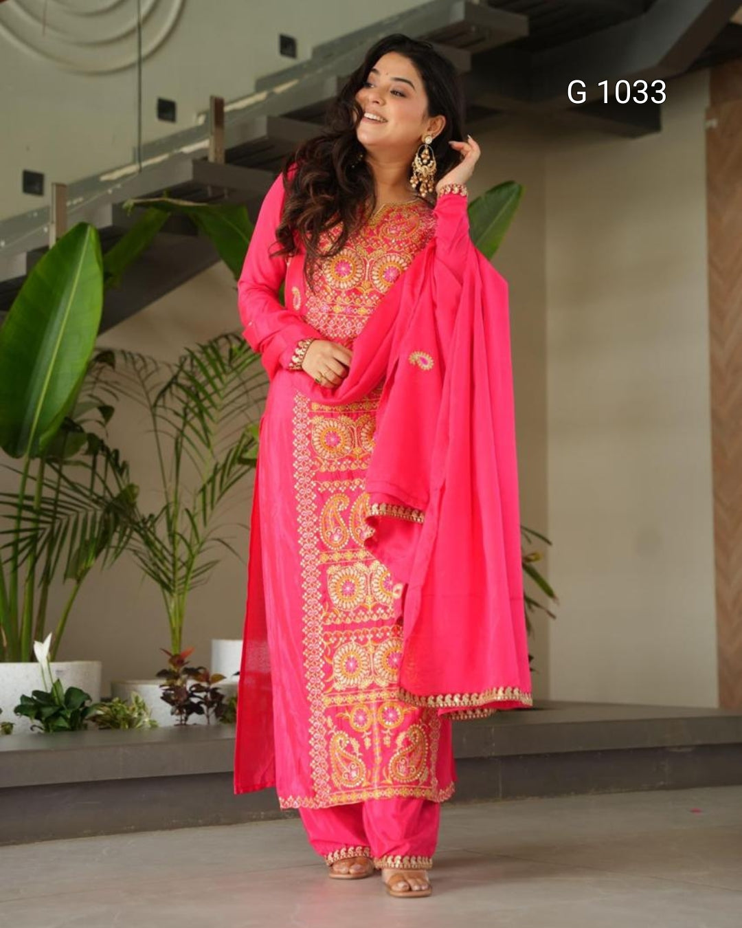 A woman modeling a bright pink traditional Indian salwar kameez suit with intricate gold and yellow embroidery, paired with a matching pink dupatta, in an indoor setting with plants.