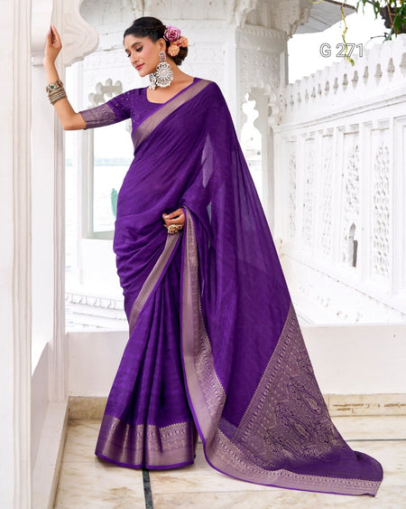 A woman models a purple saree with a matching blouse in a white architectural setting. The saree features a purple body with a contrasting lavender border and pallu, adorned with intricate metallic embroidery.
