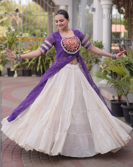 A woman modeling a traditional Indian outfit featuring a purple embroidered kurta with a matching long scarf and a tiered cream-colored flared skirt, set against an outdoor patio background with potted plants.
