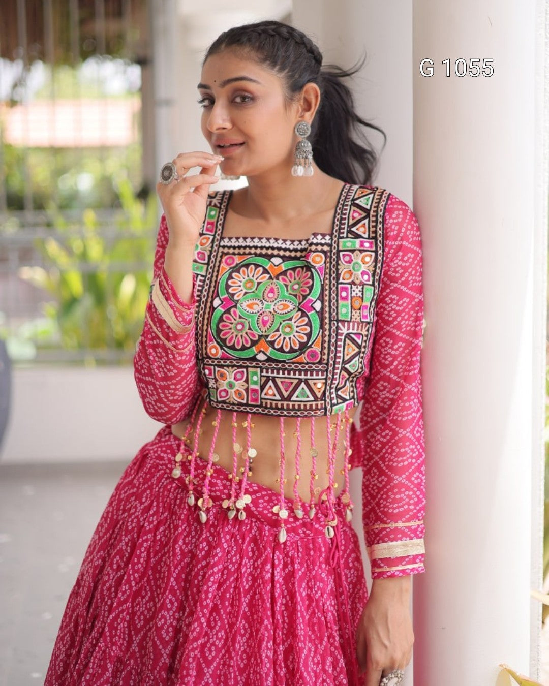 A woman wearing a bright pink embroidered traditional outfit with a patterned blouse and flared skirt, standing indoors near a white pillar. The blouse has colorful geometric and floral embroidery with dangling beaded fringe.