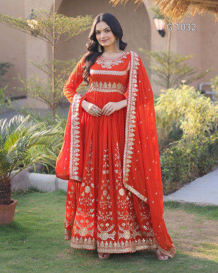 A woman models a traditional red Indian ethnic outfit with intricate gold embroidery, featuring a long kurta, matching dupatta, and a red skirt, set against an outdoor garden background.